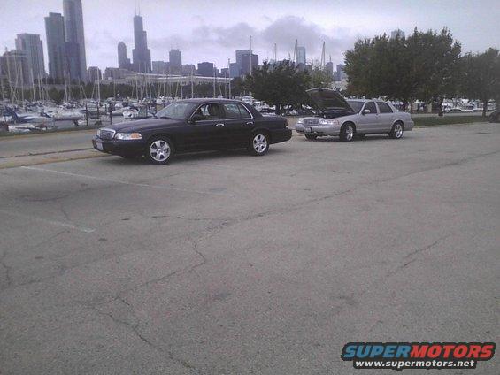 Black 2003 Ford Crown Victoria parked on asphalt, with a silver one behind it with the hood up. City skyline and marina in background
