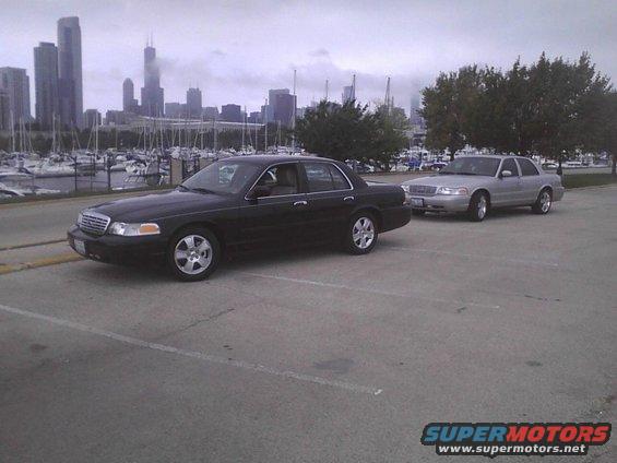Two black and silver Ford Crown Victorias parked near a marina with a city skyline in the background