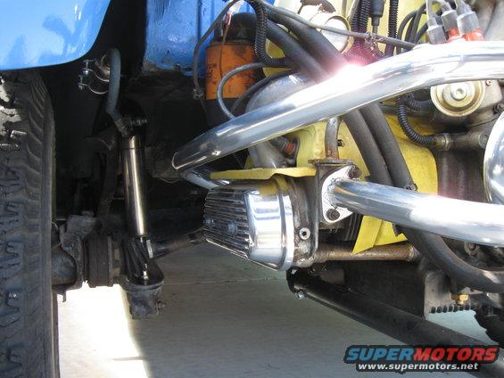 Close-up of a modified VW engine with chrome valve covers and exhaust pipes, showing suspension components and a tire