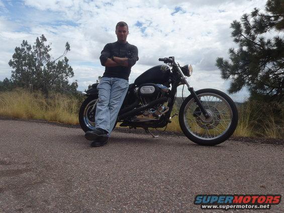 Man in jeans and dark shirt leaning on a black 2002 Harley-Davidson XL Sportster motorcycle on a paved road