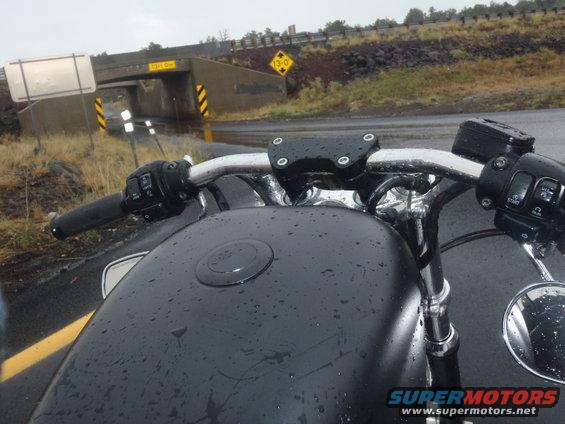 Motorcycle handlebars and black fuel tank with water droplets. Bridge and road visible in background