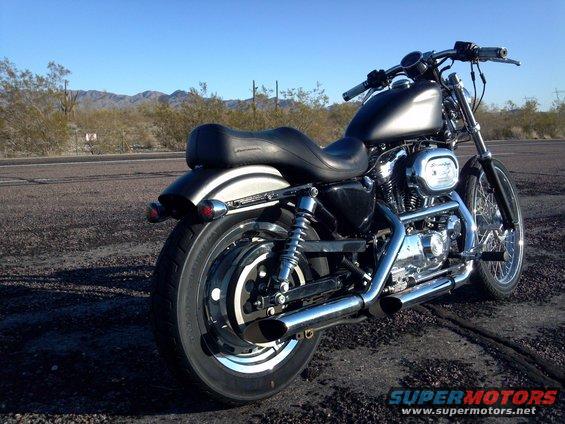 Black Harley-Davidson motorcycle parked on a road with desert landscape behind it. Shows the rear wheel, seat, and engine