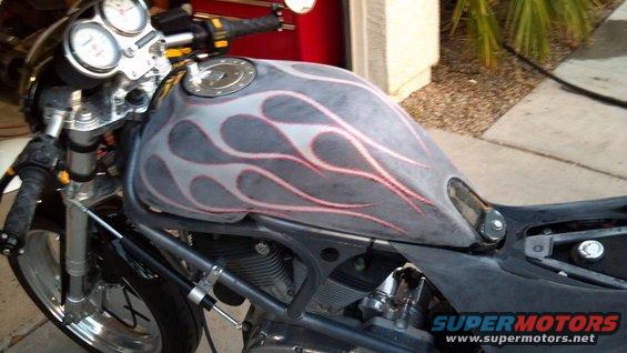Motorcycle fuel tank with gray and pink flame graphics. The front forks and part of the engine are visible