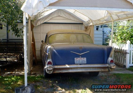 Blue 1957 sedan parked under a carport, rear view. Dusty, with chrome bumper and tail fins
