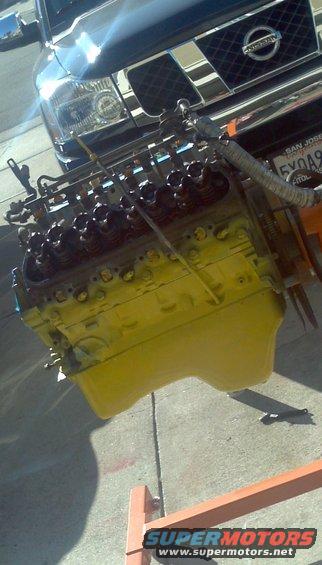 Yellow engine on an engine stand, with the front of a dark Nissan Titan truck behind it. License plate visible