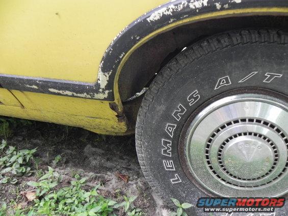 Close-up of a yellow car's rear wheel well, showing a worn tire and fender trim. Ford logo on hubcap