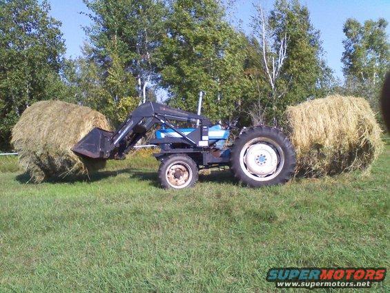 Blue tractor with a front loader, parked in a grassy field next to two large hay bales