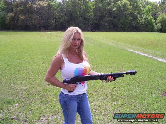 Woman in jeans and white tank top holding a black shotgun in a grassy field