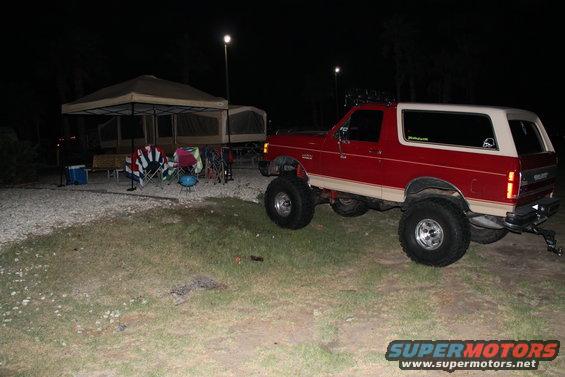 1990 Ford Bronco parked at a campsite at night, lifted suspension, red exterior with tan trim