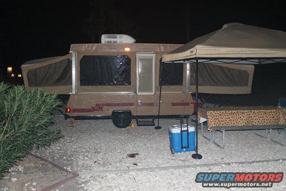 Brown pop-up camper with extended canvas sections, parked at night under a canopy
