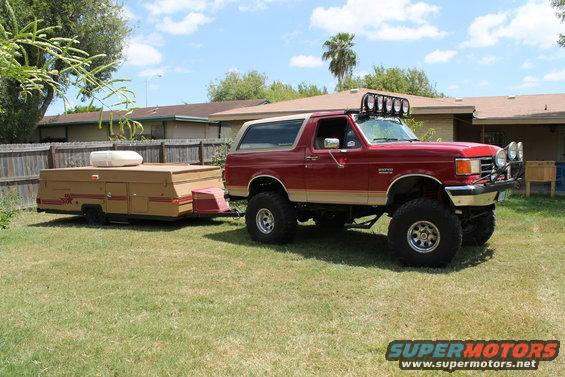 1990 Ford Bronco with lifted suspension and off-road lights, towing a tan pop-up camper