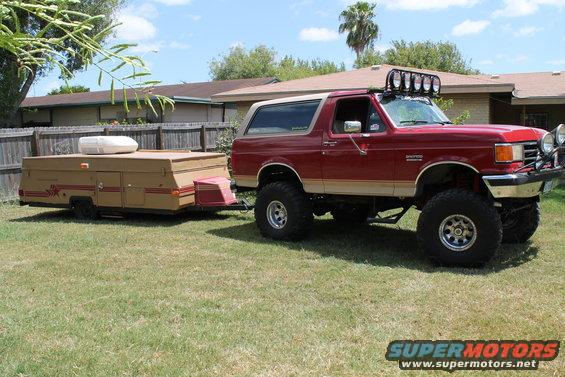 Lifted red Ford Bronco towing a tan pop-up camper in a grassy yard