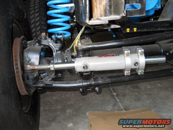Close-up of a vehicle's steering linkage with a JCR power steering assist cylinder installed. Blue coil spring visible