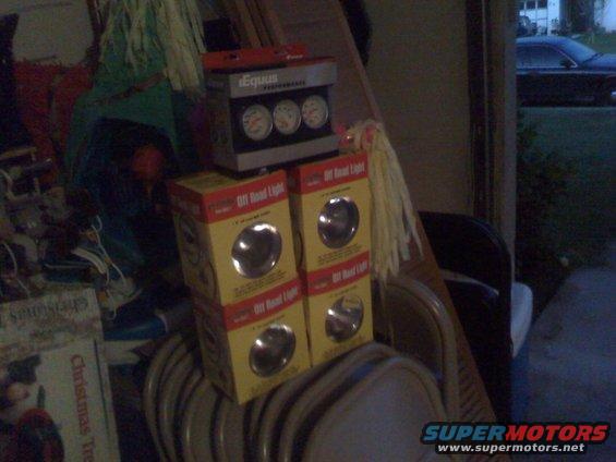 Boxes of off-road lights stacked with a gauge cluster, and a dark sedan visible in the background