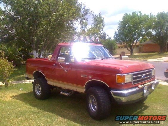 Red Ford Bronco with lift kit, off-road tires, and light bar on roof. Side view shows two-tone paint