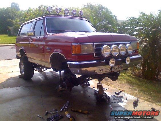 1990 Ford Bronco with front suspension removed, sitting on jack stands. Four driving lights are mounted above the bumper