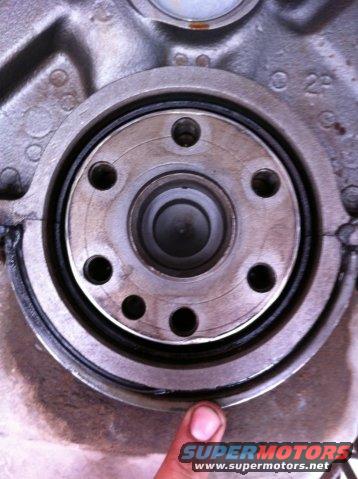 Engine crankshaft seal with a finger pointing to its edge. Text 'SUPERMOTORS' is visible