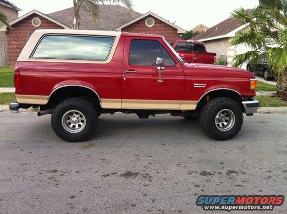 Red 1990 Ford Bronco, side view, tan top. Another red truck is visible