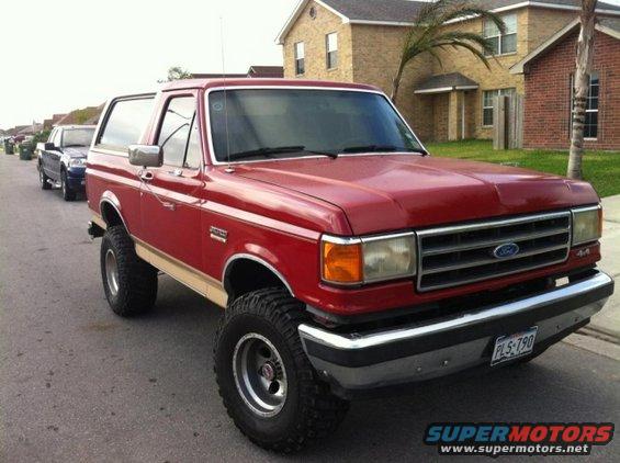 Red 1990 Ford Bronco lifted with off-road tires, parked on a residential street. Another vehicle is visible behind it