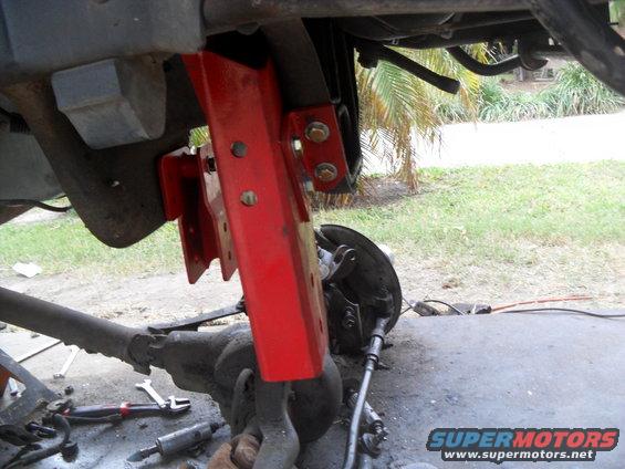 Red suspension bracket attached to a vehicle's front axle assembly, with tools on the ground