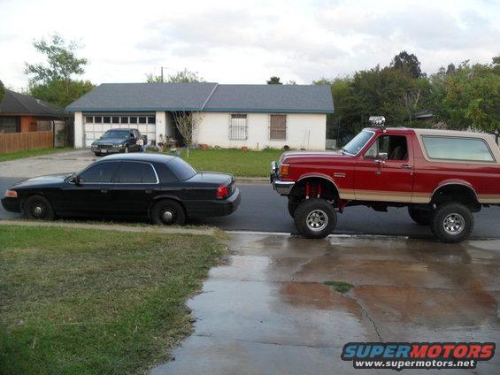 Red lifted Ford Bronco with tan top, side view. Black sedan parked on grass