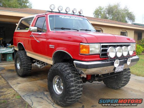 Red 1990 Ford Bronco lifted with large tires and roof-mounted lights