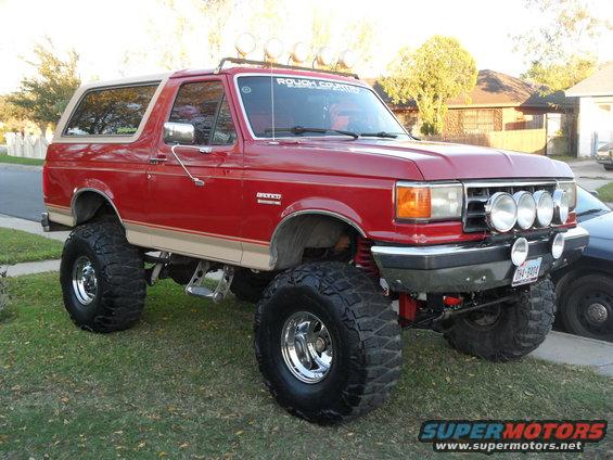Lifted red 1990 Ford Bronco with four auxiliary lights on the front bumper and roof rack