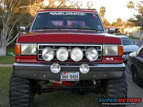 Red lifted 4x4 SUV with four large lights on grille and two smaller ones below. Has 'ROUGH COUNTRY' sticker on windshield. Texas license plate DH4-B404