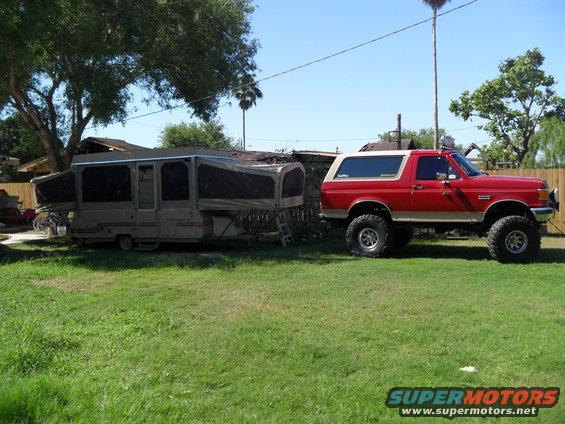 Red 1990 Ford Bronco with lifted suspension and off-road tires, parked next to a pop-up camper