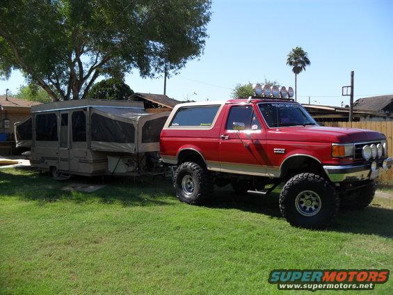 Red 1990 Ford Bronco with lifted suspension and off-road lights, towing a pop-up camper
