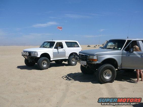 1988 Ford Bronco and a later model Ford Bronco parked on sand dunes under a clear blue sky
