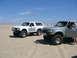 1988 Ford Bronco - Two Ford Broncos, one from 1988, are parked on a sandy beach with a checkered flag in the sky.