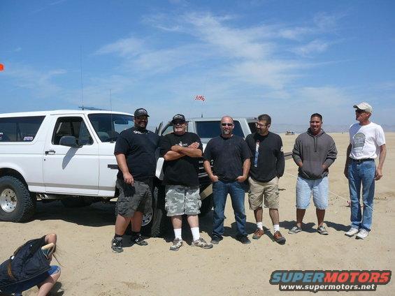 White Ford Bronco parked on sand with five guys standing next to it. Another SUV is behind it