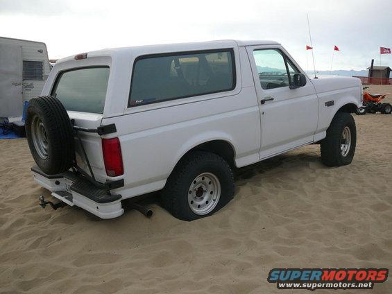 White 1992 Ford Bronco stuck in sand, spare tire on rear door, trailer and ATV in background