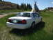 2006 Ford Crown Victoria - A white sedan is parked in a grassy field, with a teepee and a house visible in the background.