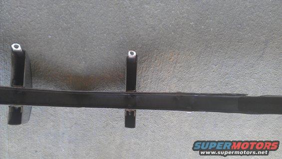 Black metal car part with two vertical supports, lying on a concrete surface. Supermotors.net logo in lower right