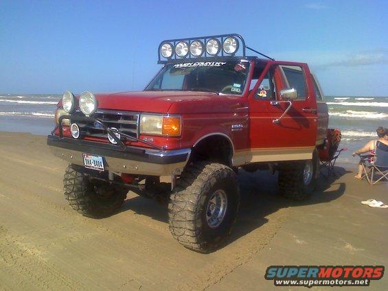 Lifted red Ford Bronco with off-road lights parked on a sandy beach near the ocean