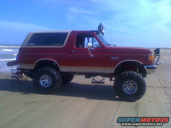 Red lifted 1990 Ford Bronco on beach with aftermarket lights