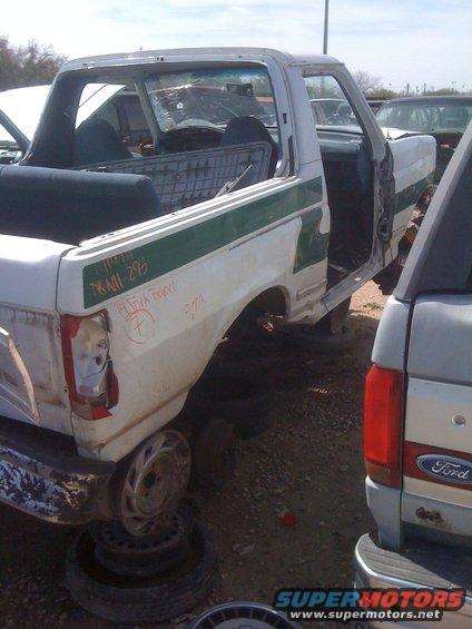 White pickup truck with green stripe, missing doors and wheels, parked in junkyard. Another Ford truck is partially visible to the right