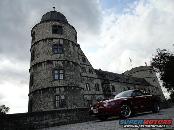 Red coupe with aftermarket wheels parked on a cobblestone drive in front of a stone castle