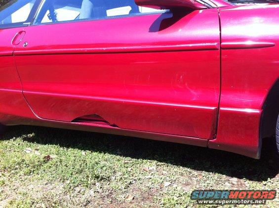 Red car door with rust damage on the lower rocker panel, viewed from the side