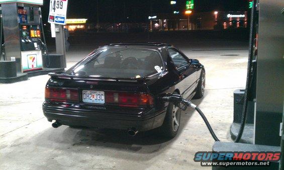 Black 1988 Mazda RX-7 with a gas nozzle in the fuel door, parked at a gas station at night