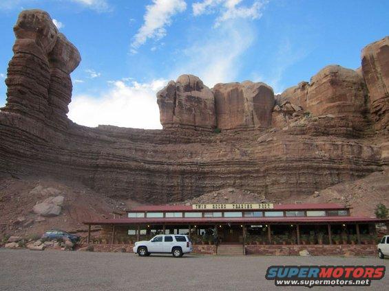 White SUV parked in front of a building with red rock formations. A dark sedan is also visible