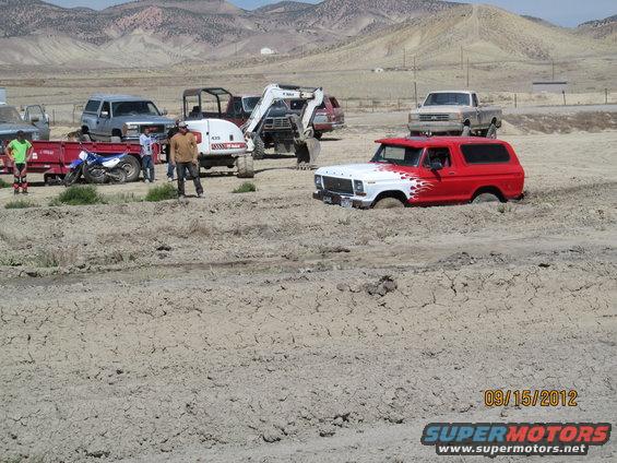 Red and white Ford Bronco stuck in mud, with other vehicles and people around