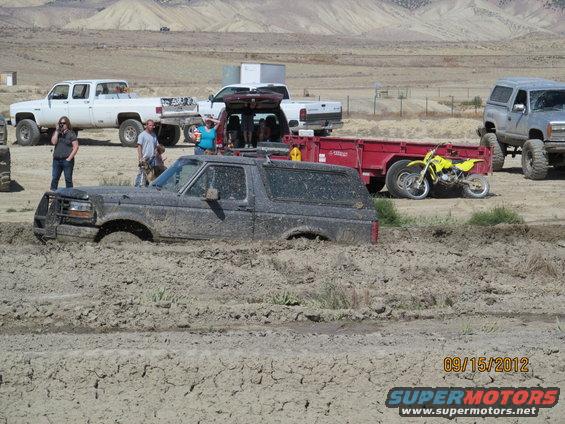 Mud-covered 1995 Ford Bronco stuck in deep mud, with other trucks and a dirt bike visible in the background