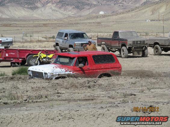 Red and white 1978 Ford Bronco stuck in mud, with other trucks and a motorcycle nearby