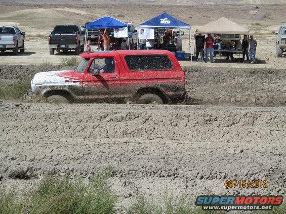 Red 1979 Ford Bronco driving through a mud pit with other vehicles and tents in the background