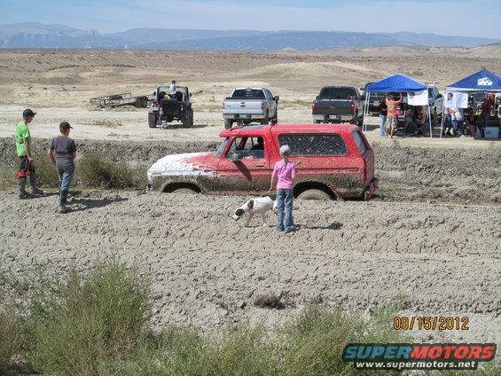 Red Ford Bronco stuck deep in mud, a person and dog stand nearby. Other vehicles and tents are visible in the background. Date stamp reads 09/15/2012
