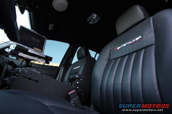 2004 Ford Crown Victoria police car interior showing front seats, console with laptop, and 'GHOSTALKER' embroidery on passenger seat
