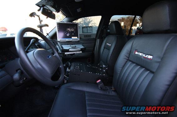 Interior of a police car showing a steering wheel, laptop with 'POLICE' on screen, and custom seats with 'GHOSTALKER' logo